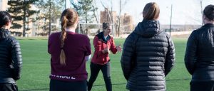 Image of Coach Bert instructing a group of college players on a field in Colorado. Bert is leaning forward, smiling, and gesturing with open hands.