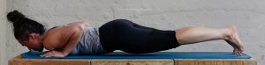 Image of Coach Kira, face-down on a mat in front of a white wall. Kira is performing a core exercise with her hands under her shoulders, elbows bent, and legs hovering off the ground with her toes dug into the mat.