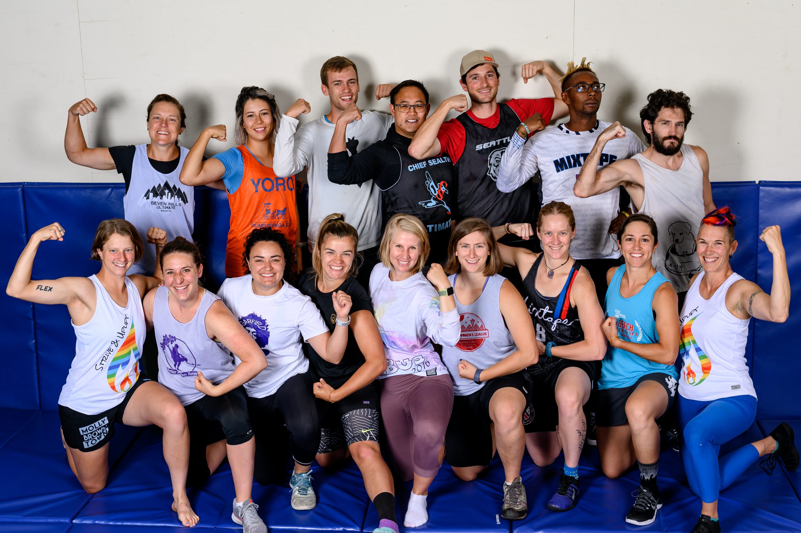 Image of a group of ultimate players in two rows wearing team jerseys with coaches Bert and Ren. Everyone is flexing their arms and smiling or making fierce expressions.