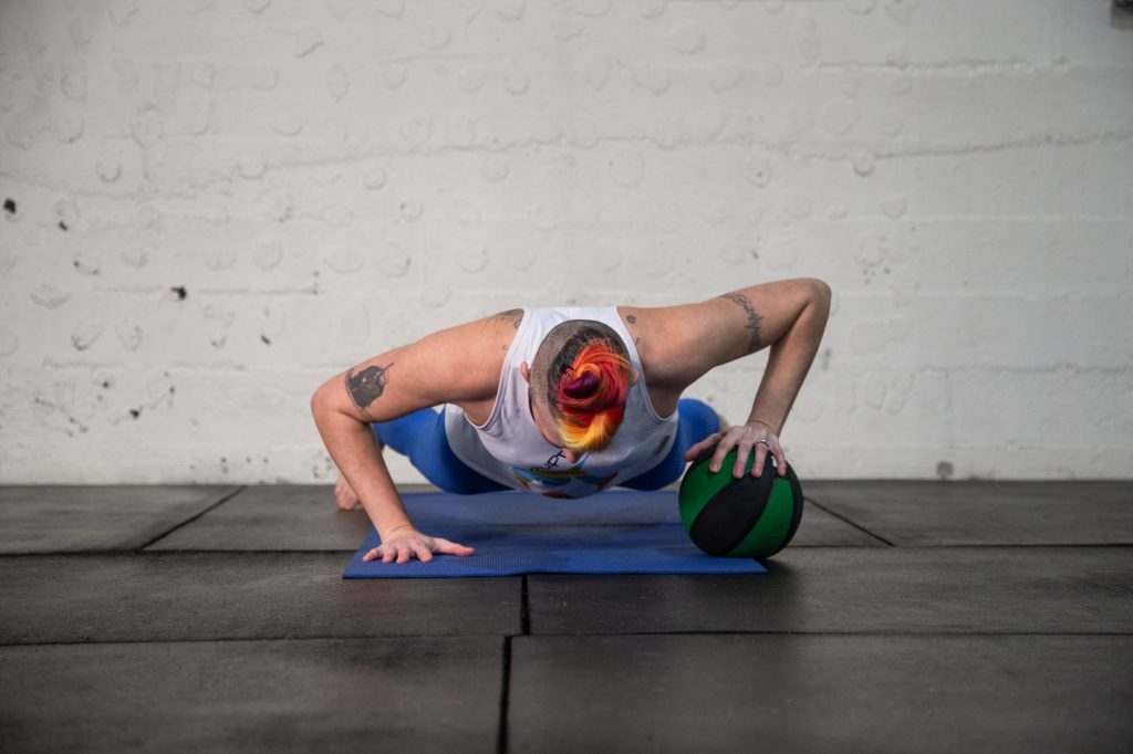 Image of Ren performing a medicine ball pushup on a blue yoga mat on a black floor in front of a white wall.