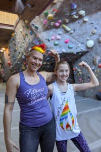 Image of Ren and their pre-teen daughter smiling and flexing their left arms inside a bouldering gym. Ren is wearing a purple tank that reads "Fitness for all Bodies" and their daughter is wearing a white tank with Strive & Uplift flame logo in rainbow stripes.