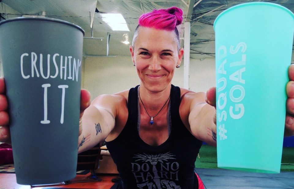Image of Ren standing in the gym wearing a black tank top, smirking at the camera while holding forward cups in each hand that say "Crushin' It" and "Squad Goals". Ren has a fresh pink dye on her bunned up hair.