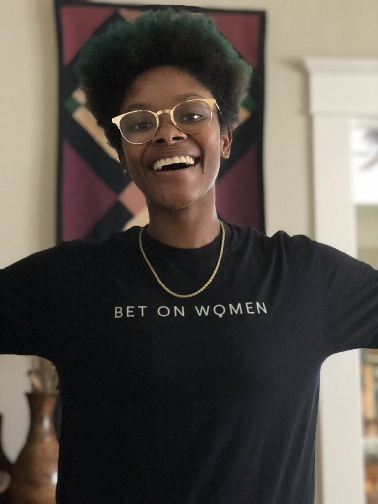 Anraya Palmer, smiles with her arms wide, wearing a "Bet on Women" shirt.