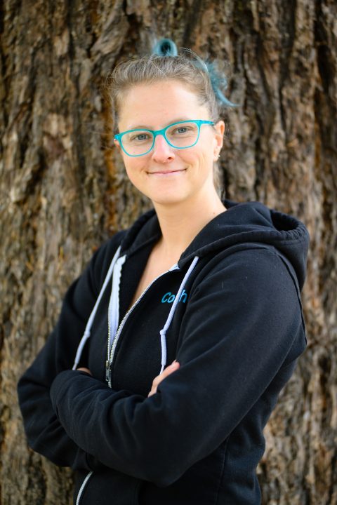 Bert Cherry posed in front of a tree trunk and wearing a black zip-up sweatshirt. Bert is a white person (she/they pronouns) with brightly colored hair and bright blue glasses.
