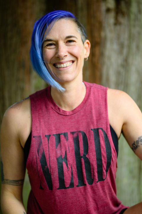 Ren Caldwell smiling, posed in front of a tree trunk in a red tank that reads "NERD". Ren is a white person (they/she pronouns) with visible tattoos and brightly colored hair.